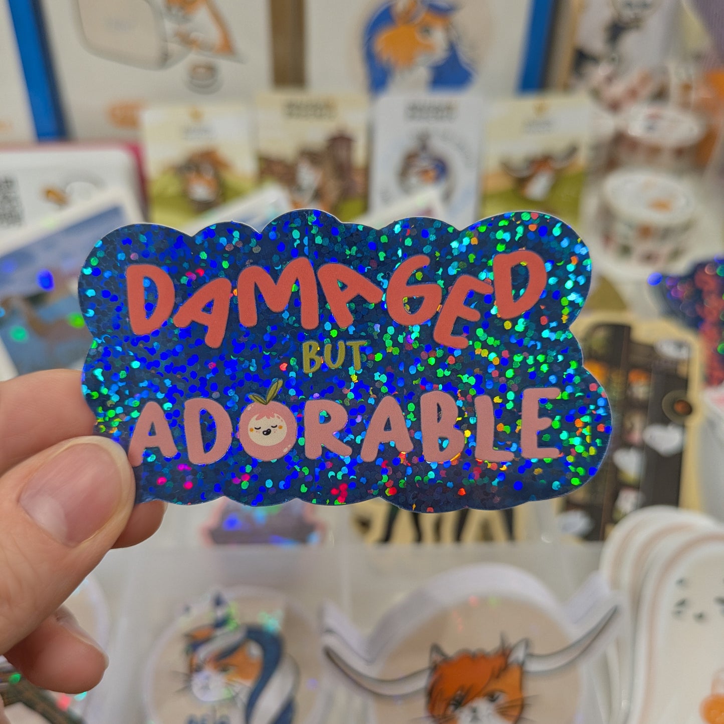Holographic sticker with 'Damaged but Adorable' text held in front of a store display.